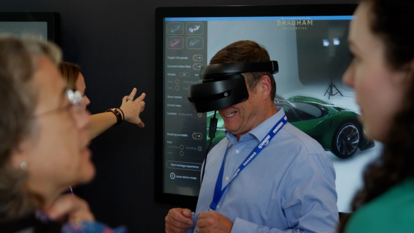 A man in a shirt smiling whilst wearing an VR headset in front of a screen showing a virtual sports car customiser.