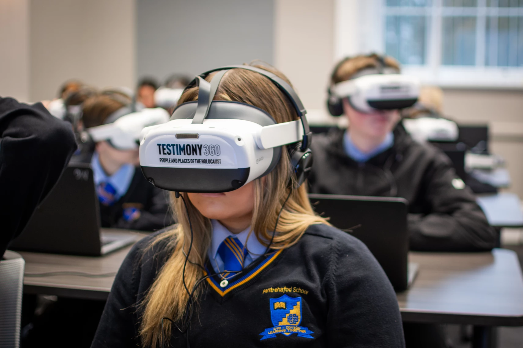 A schoolgirl in a classroom with other children sits with a VR headset on her head, with the words 'Testimony360' on it.
